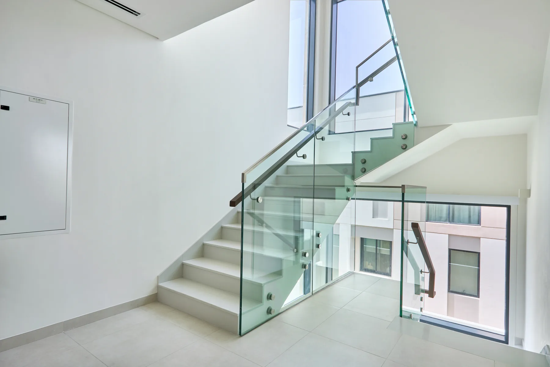 Una escalera interior luminosa tiene escalones blancos, barandillas de vidrio y ventanas grandes.