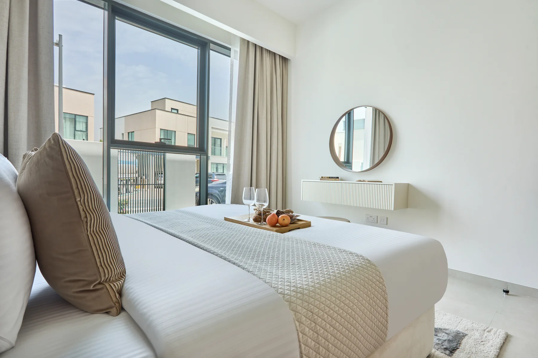 Un dormitorio luminoso con una cama junto a grandes ventanas y un espejo redondo.