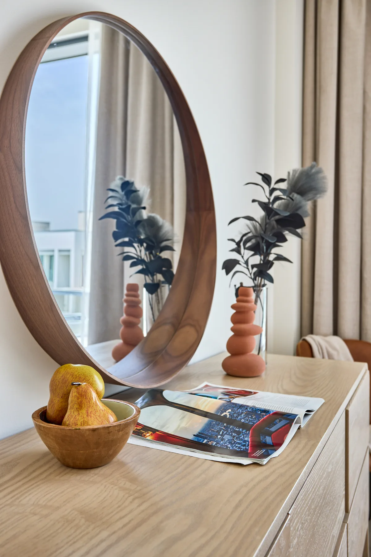 Una cómoda de dormitorio sostiene un espejo ovalado, un jarrón con flores y un cuenco de fruta.