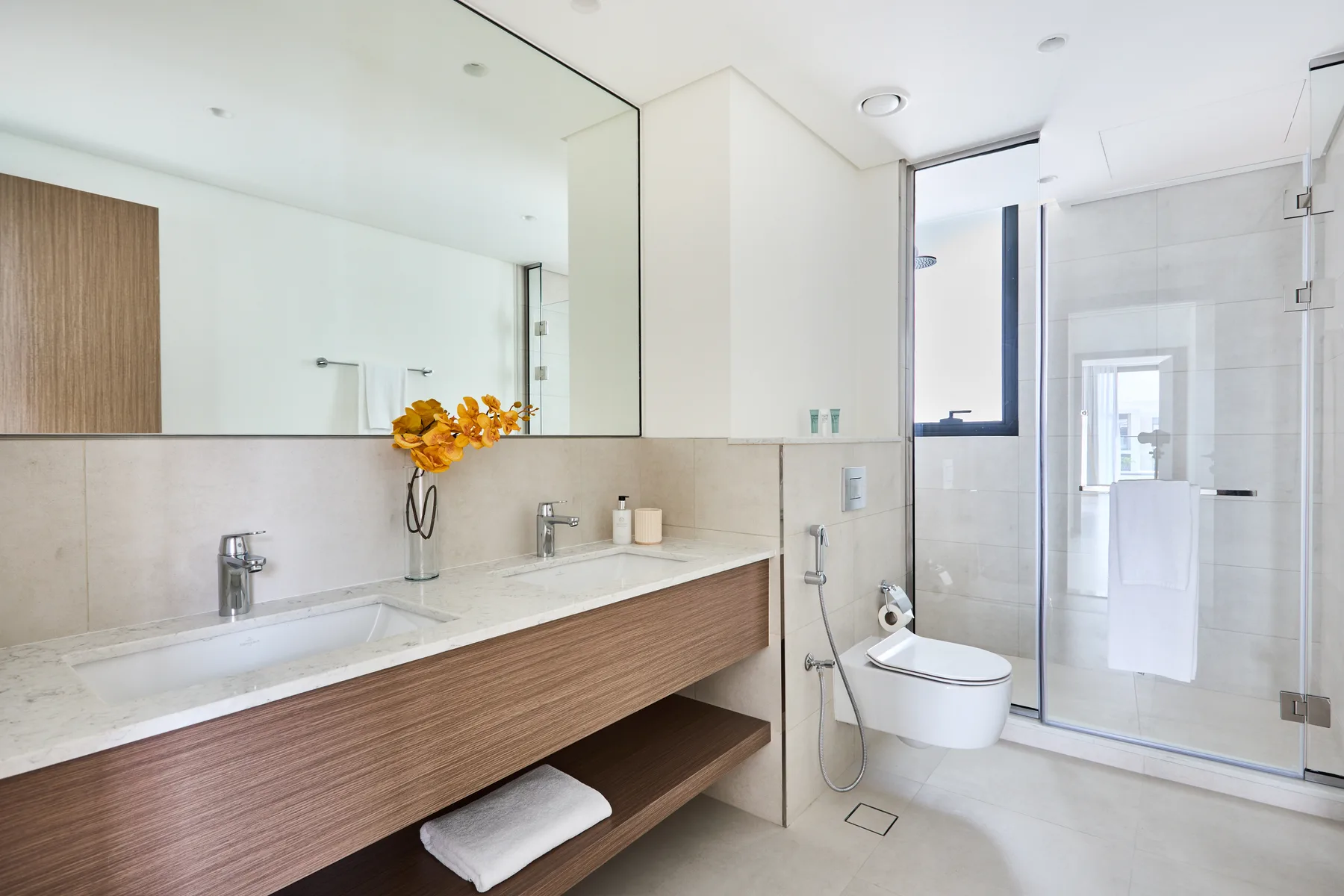 Un baño luminoso con un tocador largo, flores y una ducha con mampara de vidrio.
