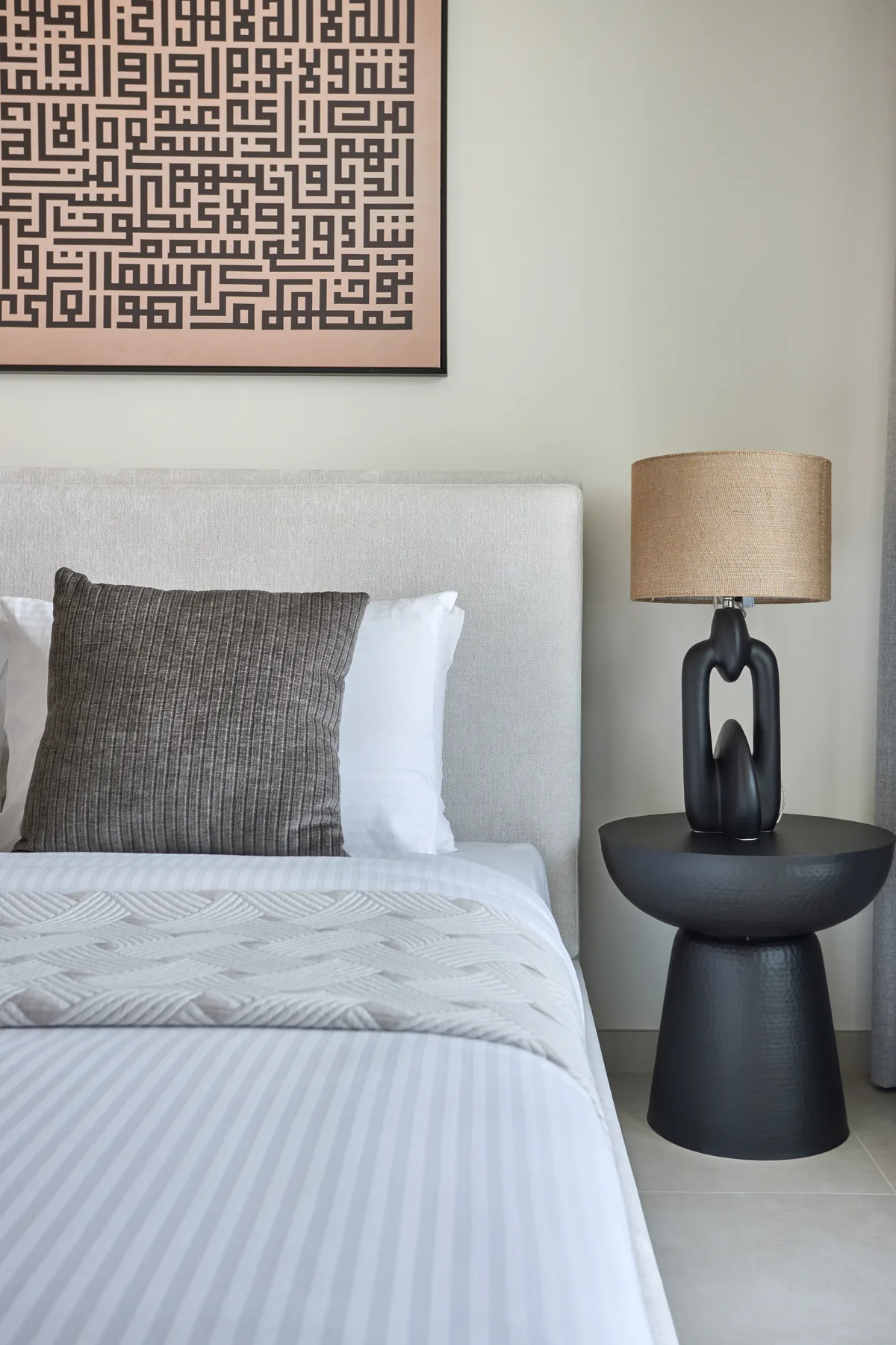 Una cama bien arreglada con varias almohadas está junto a una lámpara de mesa escultural.