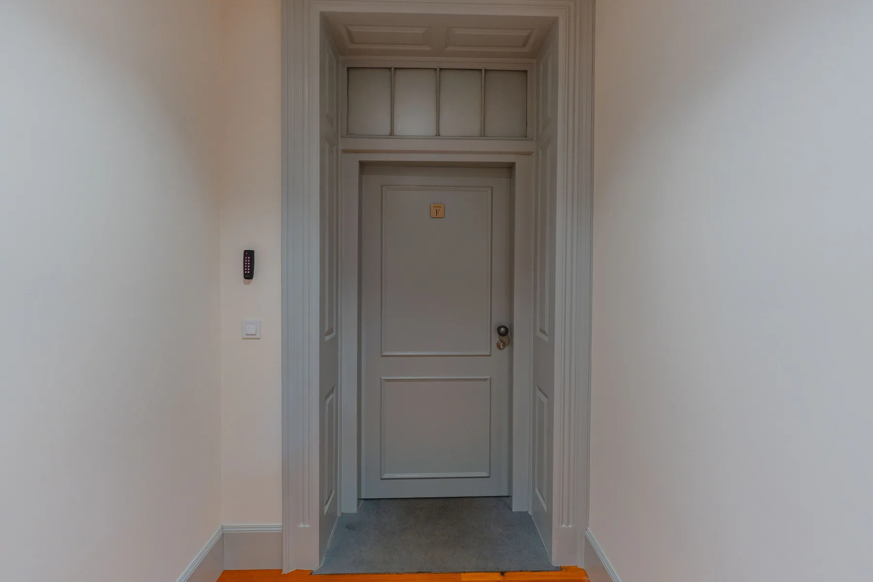 Un pasillo conduce a una puerta panelada con un pequeño control en la pared.