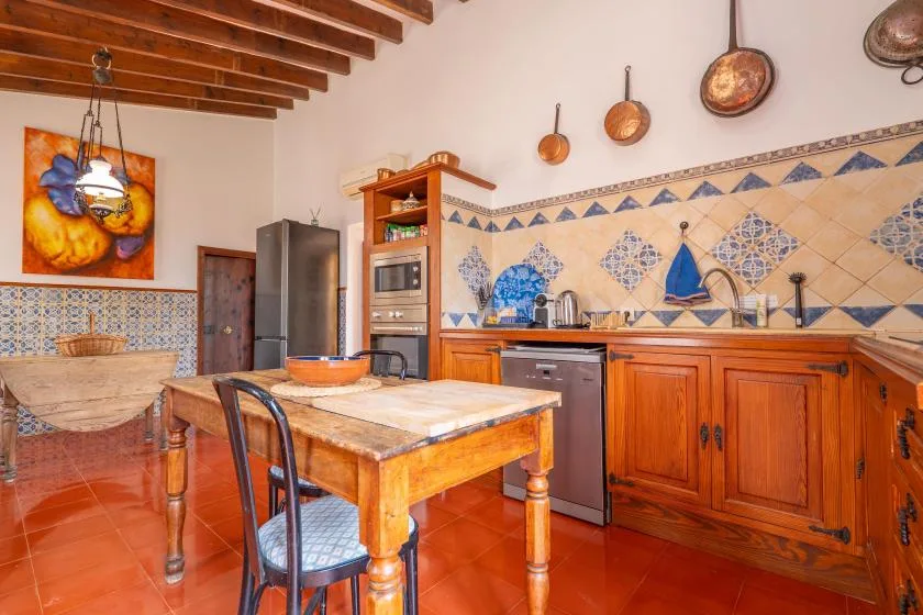 A tiled kitchen with wooden cabinets, a central table, and exposed ceiling beams.