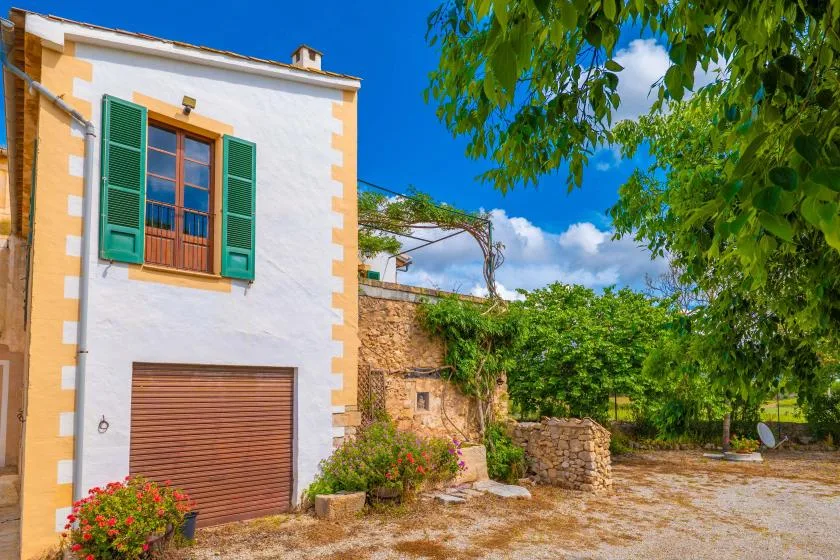 A white building with green shutters stands beside a stone wall in a sunny courtyard.