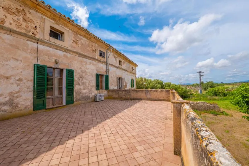 A tiled terrace runs alongside a stone building, with green shutters and open countryside beyond.