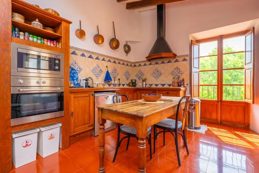 A warm kitchen with a wooden dining table and a large sunlit window.