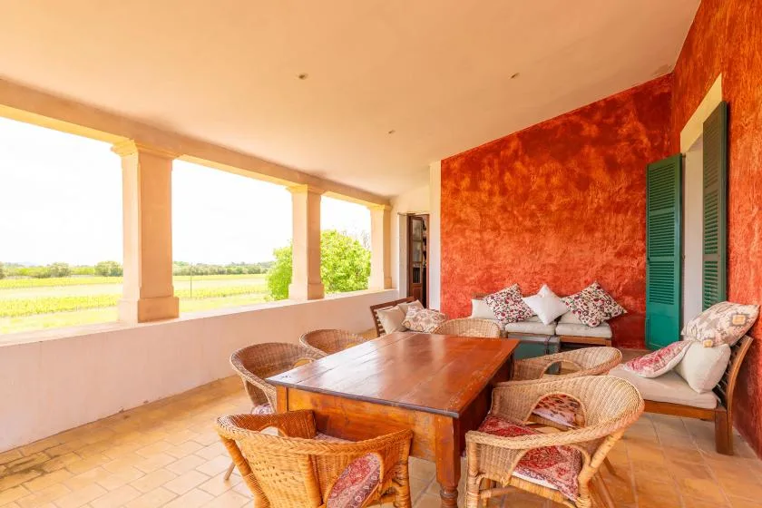 A covered patio has wicker seating around a wooden table, with open views of fields.