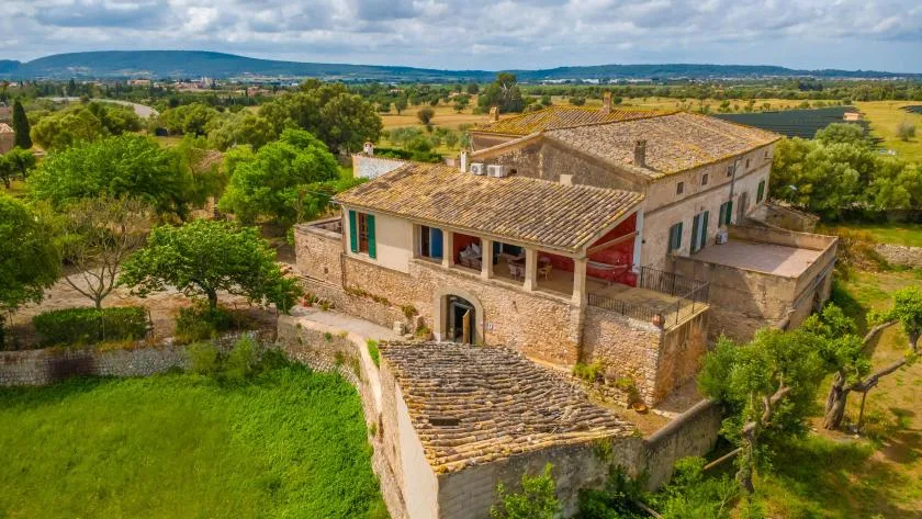 A stone country house with tiled roofs sits among green fields and trees.