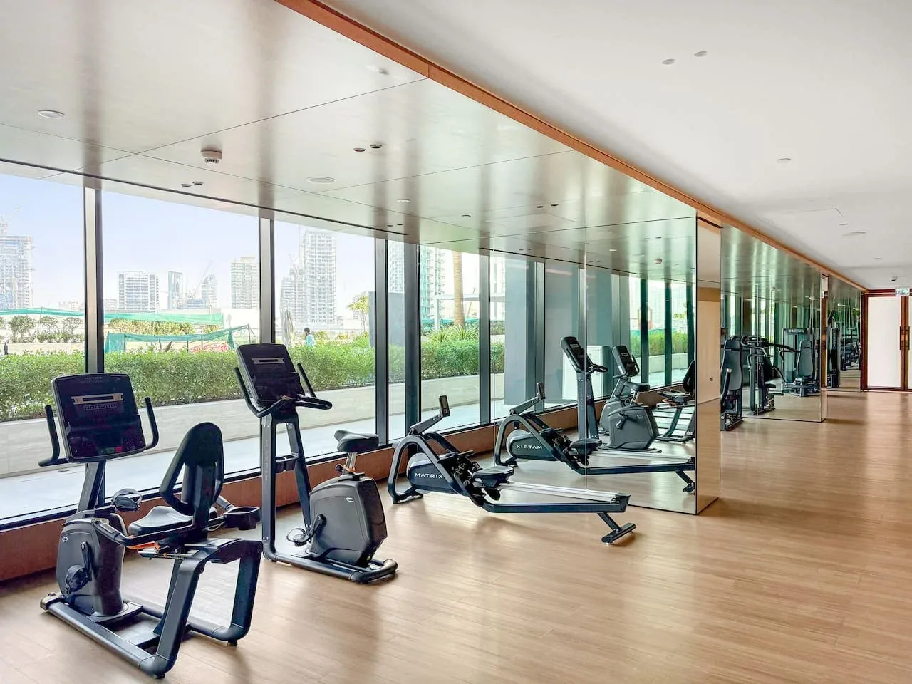 Un gimnasio con máquinas de ejercicio junto a ventanales con vistas a vegetación y edificios.