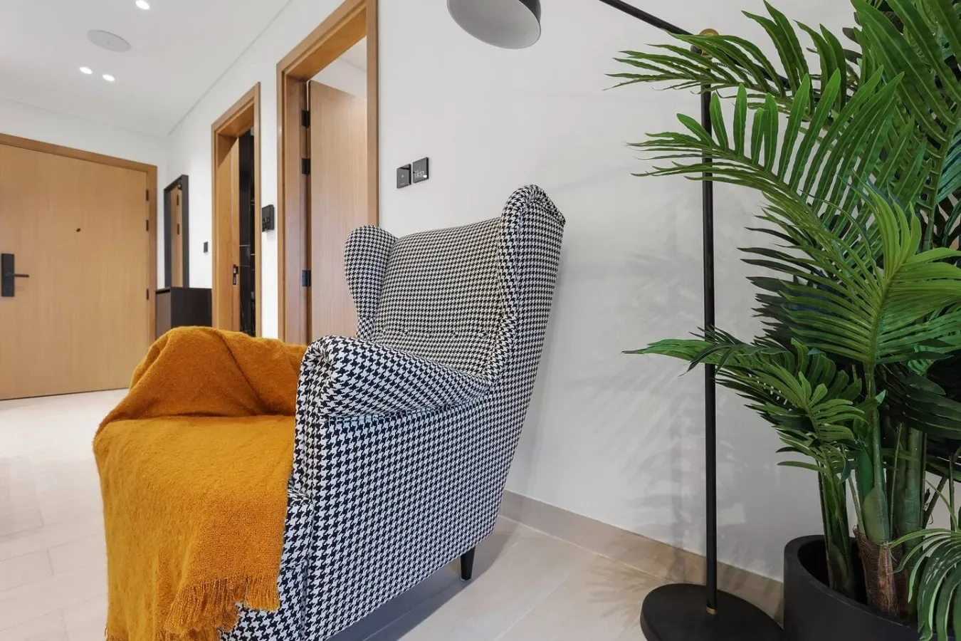Un pasillo luminoso tiene un sillón estampado, una manta mostaza y una palmera en maceta.