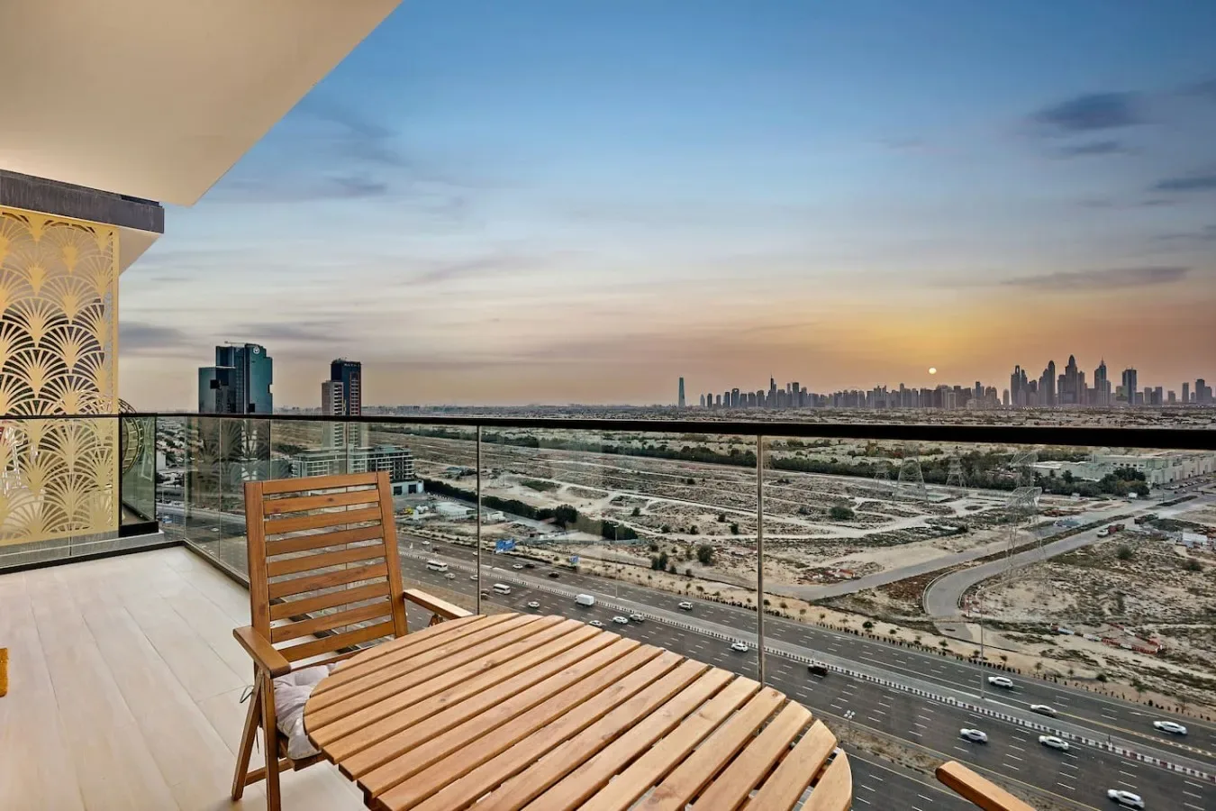 Un balcón da a un perfil urbano al atardecer, con asientos de madera junto a la barandilla de vidrio.