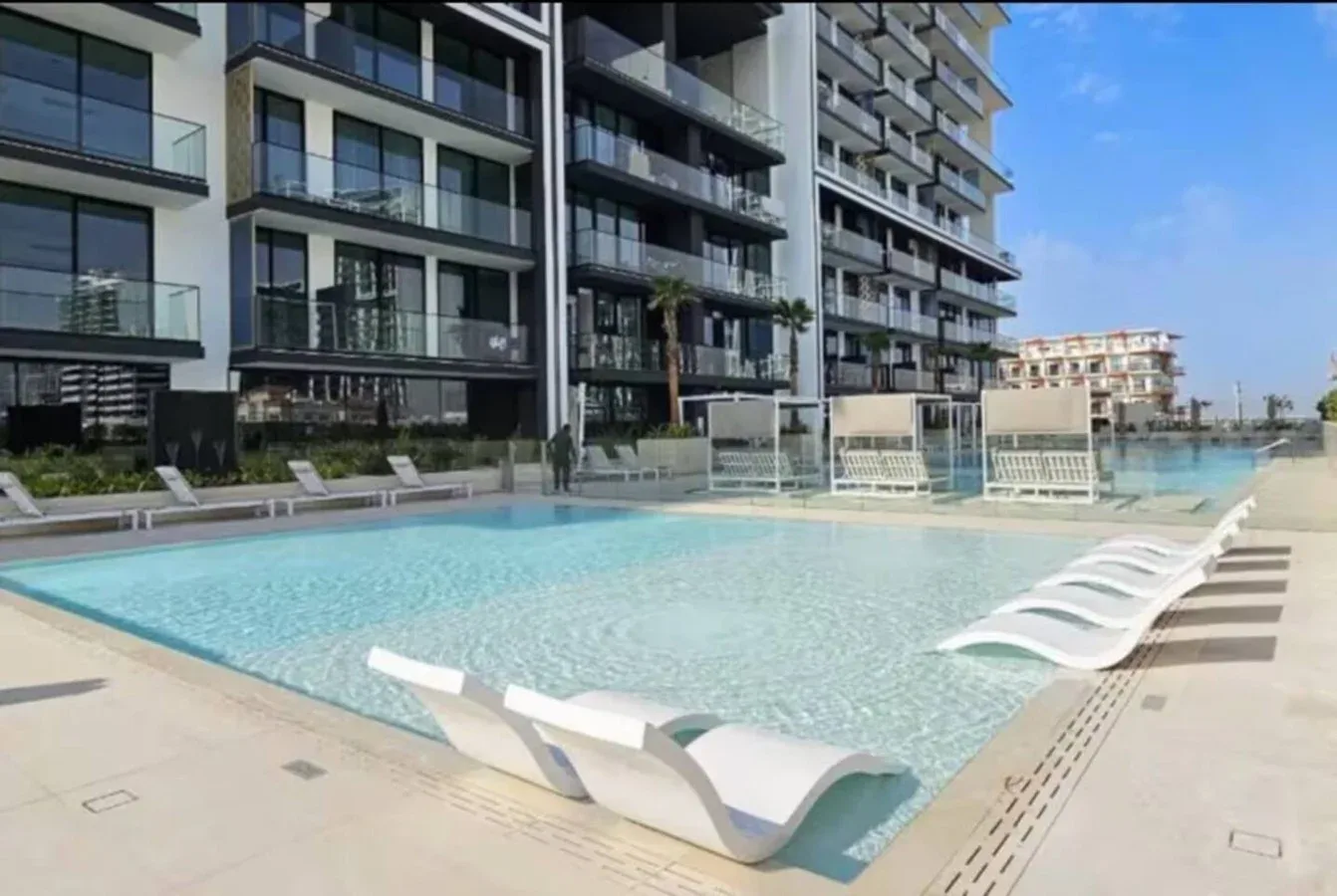 Una piscina al aire libre junto a un edificio de apartamentos moderno, con tumbonas en la terraza.