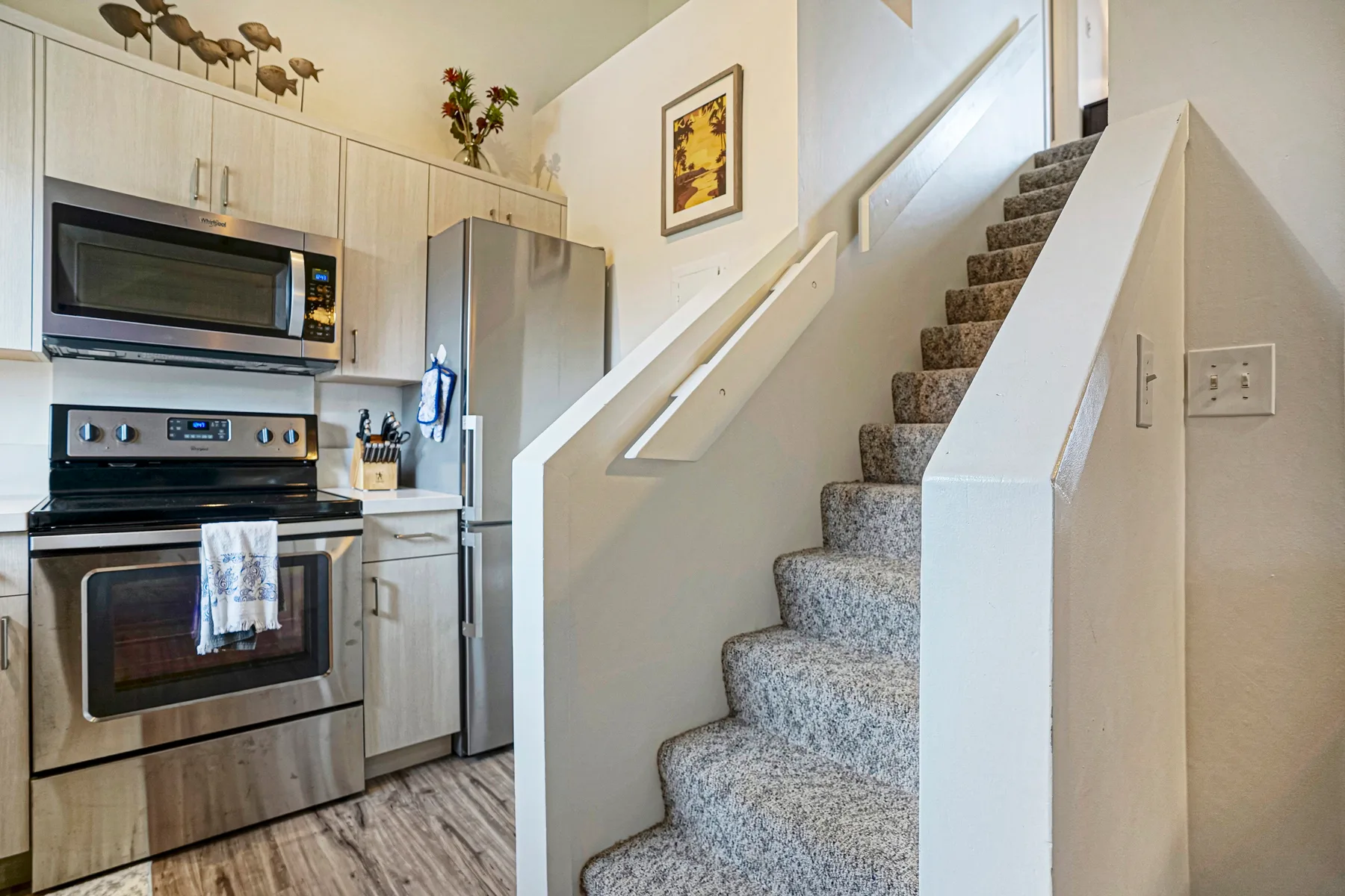 A kitchen beside a carpeted staircase, with stainless steel appliances and light cabinetry.