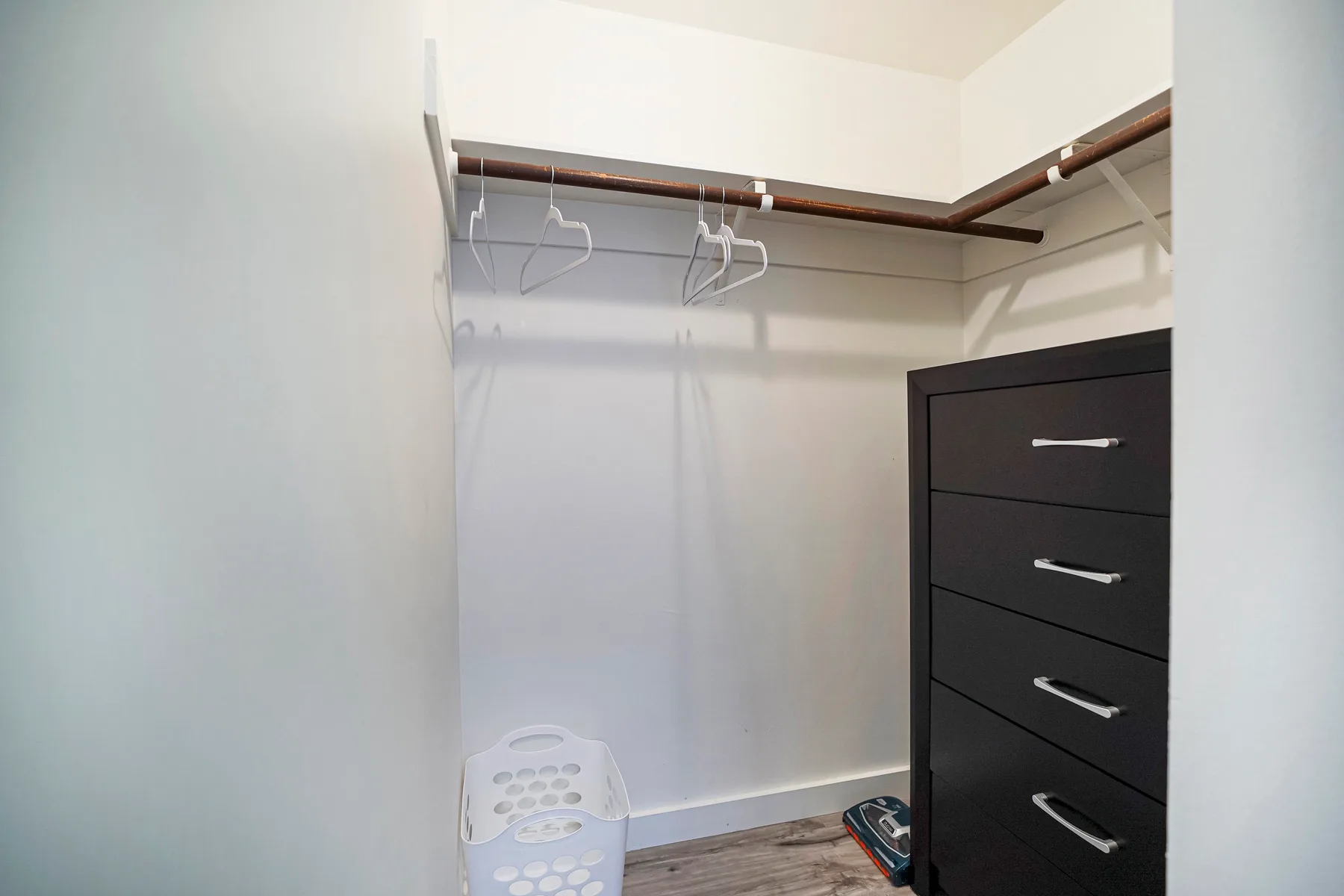 An open closet with hanging rods, hangers, and a dark chest of drawers.