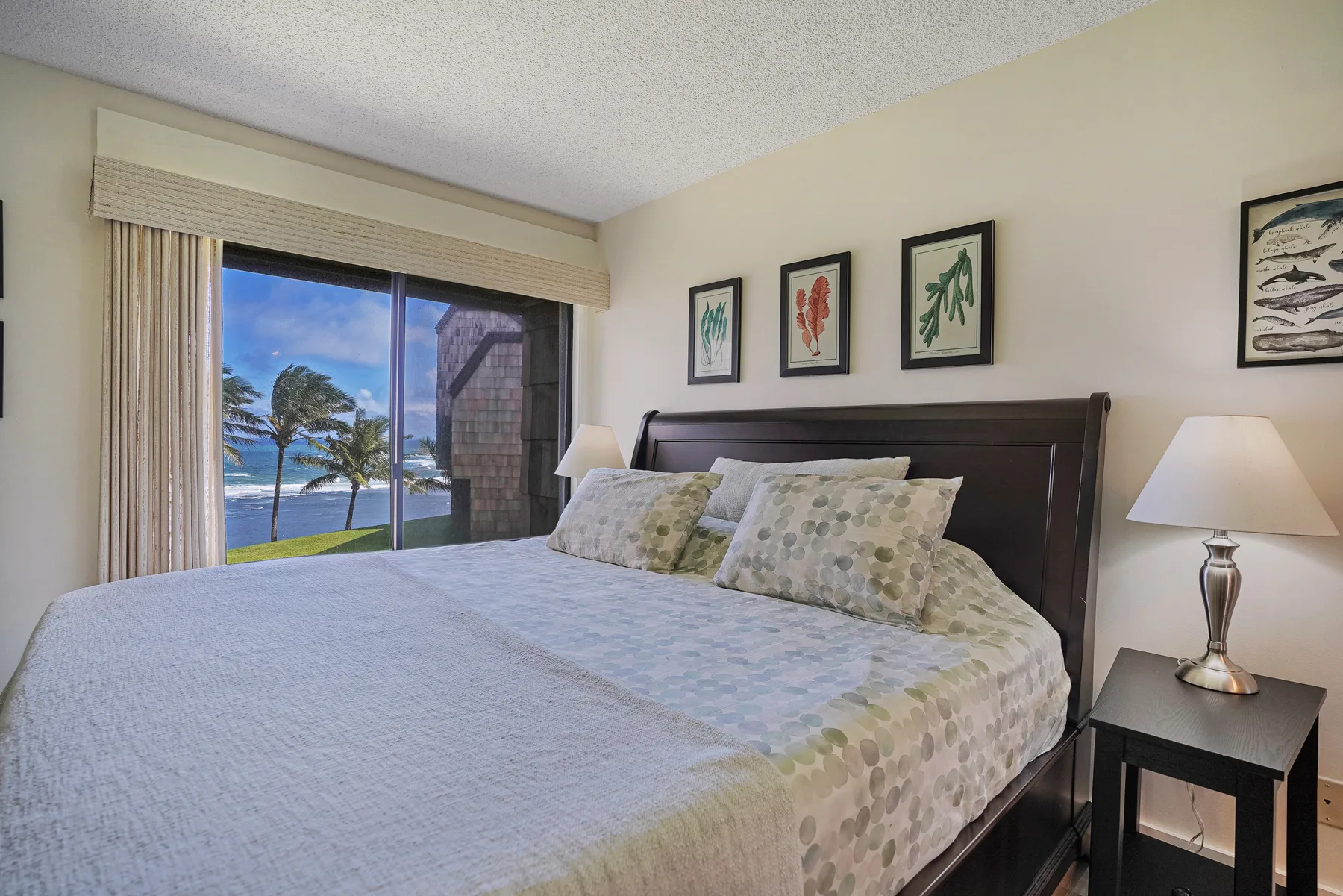 A bedroom with a large bed, framed artwork, and a window overlooking palm trees.