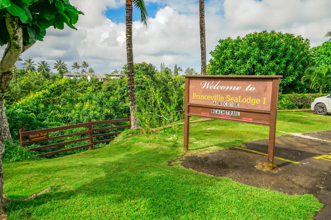 A wooden welcome sign stands beside a grassy entrance surrounded by tropical greenery.