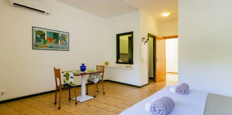 Un dormitorio con una cama, suelo de baldosas y una puerta abierta a un espacio contiguo iluminado.