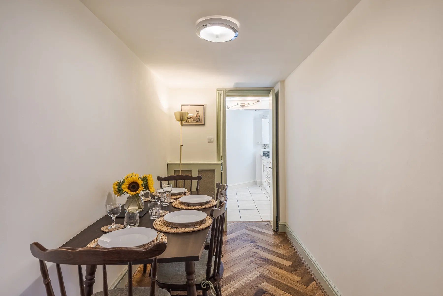 A narrow dining area with a set table leads to a bright tiled room.