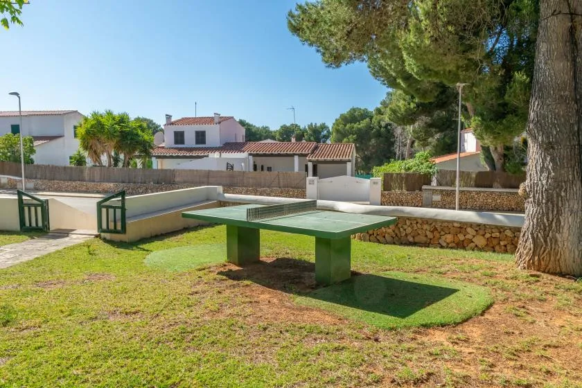 An outdoor table tennis area sits on a grassy lawn beside trees and low stone walls.
