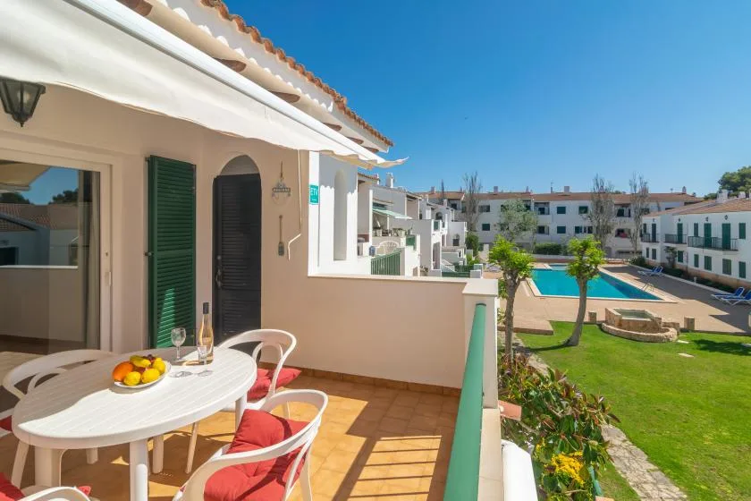 A sunlit balcony with outdoor seating overlooks a swimming pool and landscaped grounds.