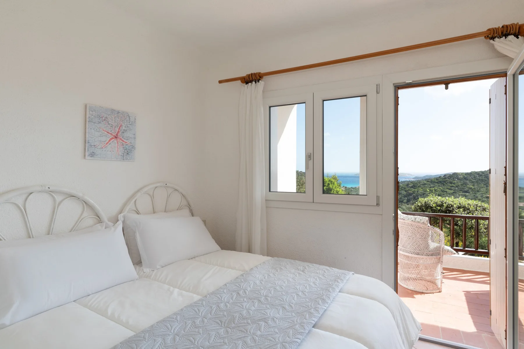 Un dormitorio luminoso con ropa de cama blanca y una puerta abierta al balcón con vistas a la vegetación.