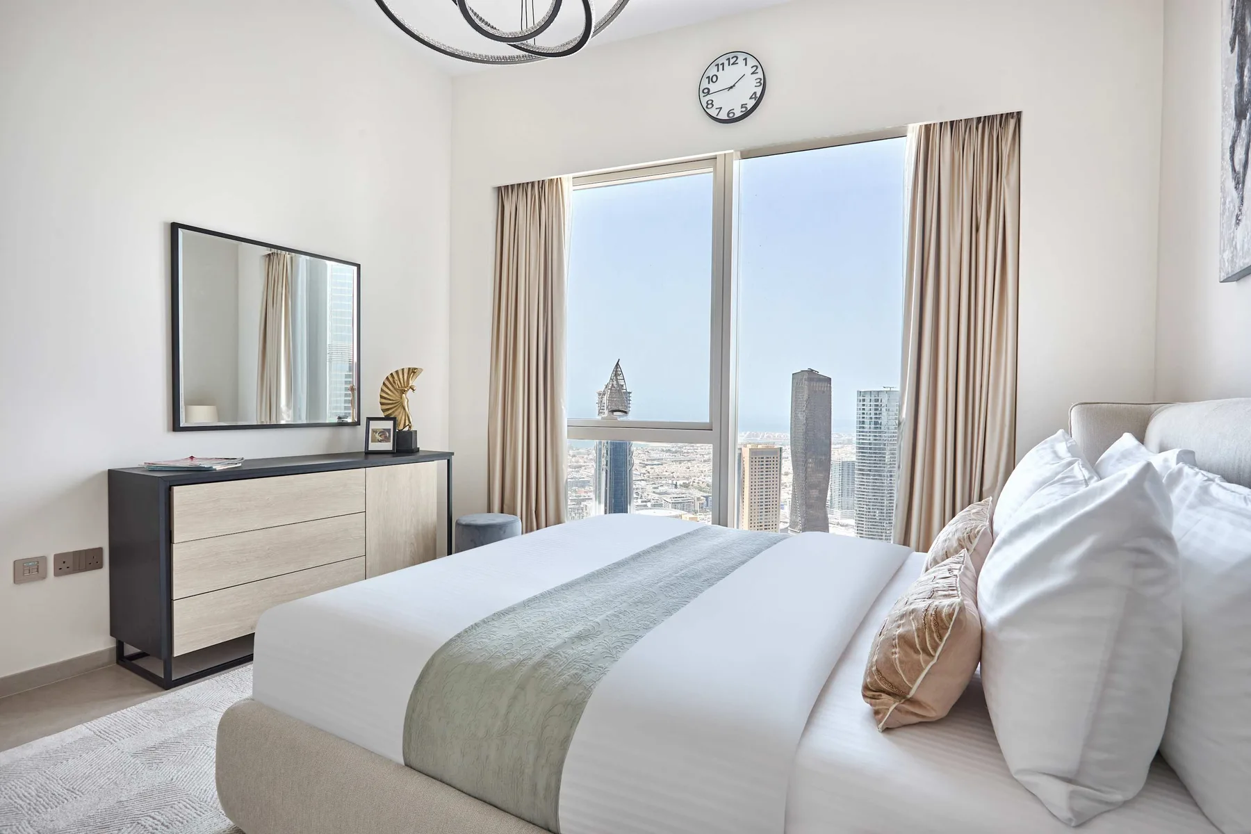 Un dormitorio luminoso con una cama tendida junto a grandes ventanas con vistas a edificios de la ciudad.