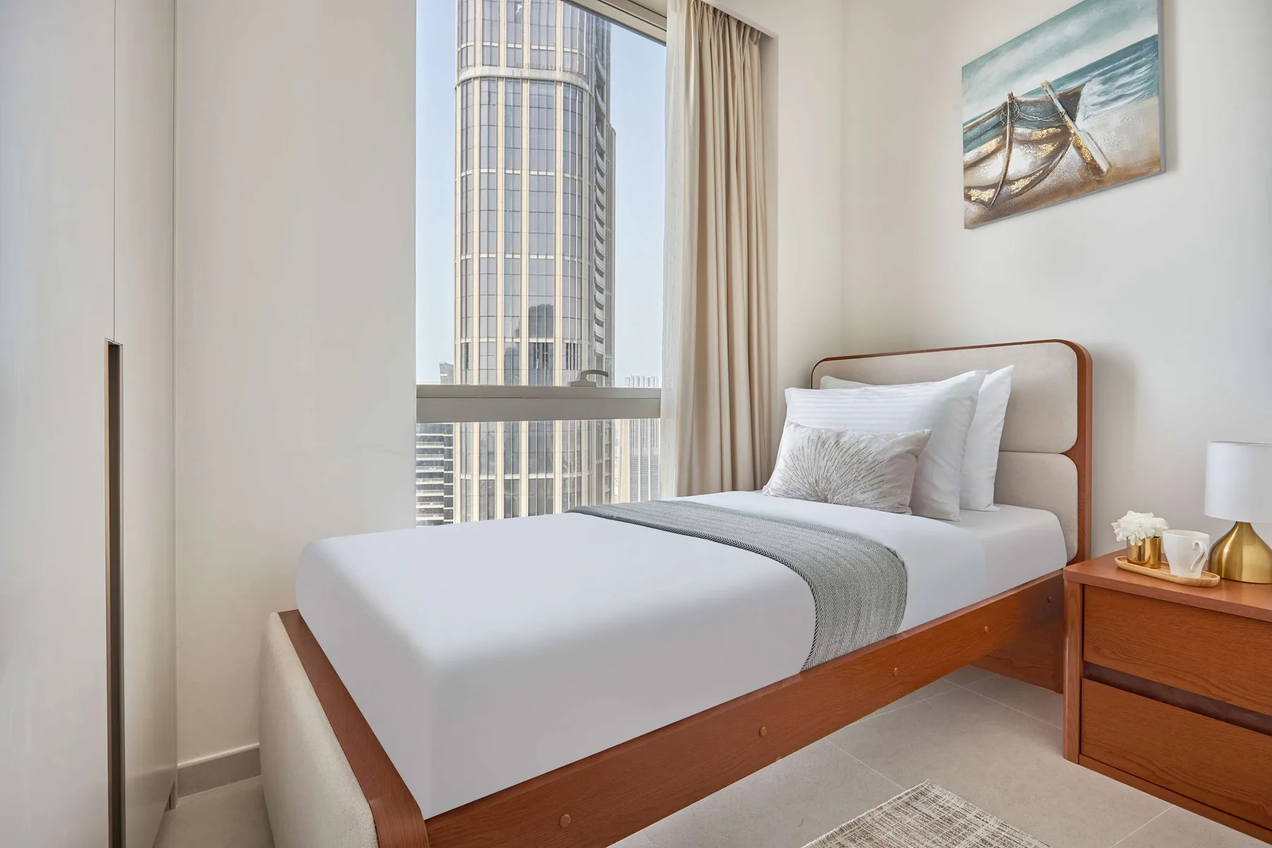 Un dormitorio con una cama bien tendida junto a una gran ventana con vistas a edificios cercanos.