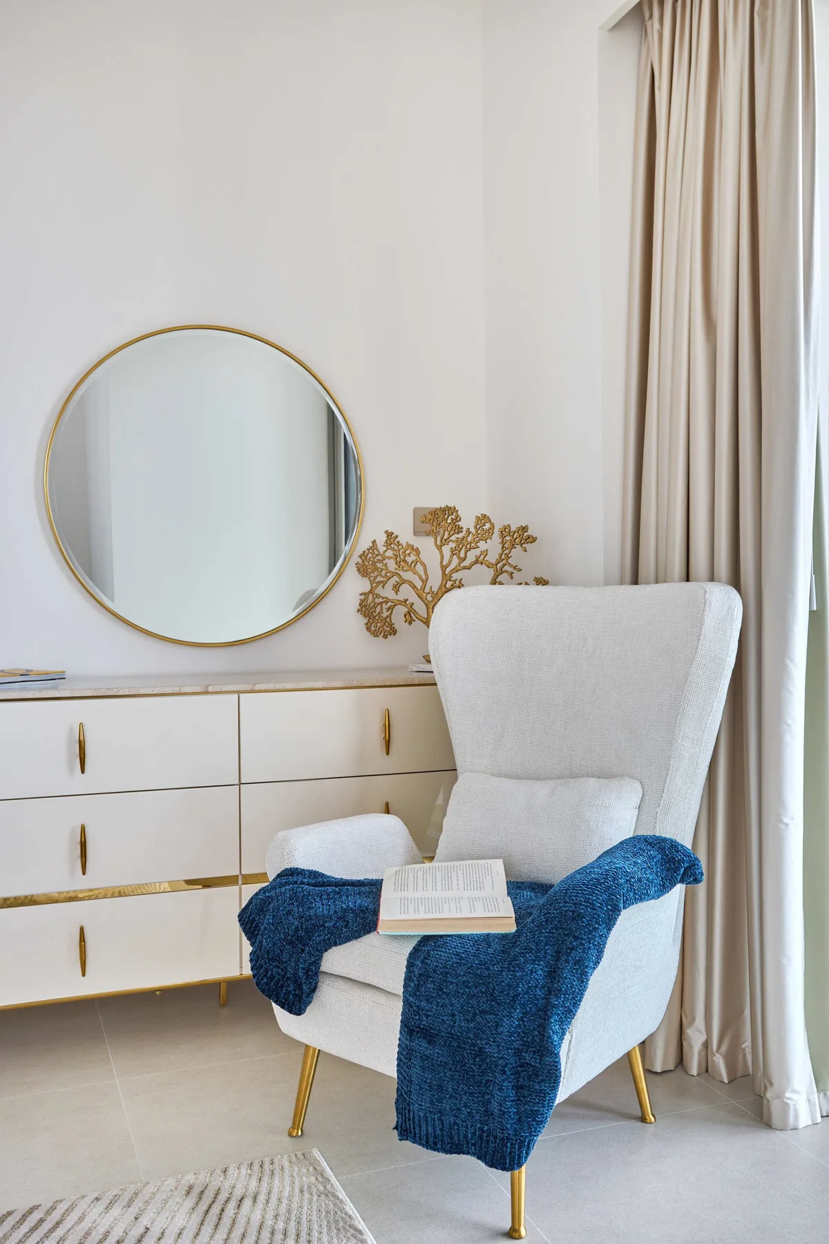 Un sillón blanco con una manta azul está junto a una cómoda y un espejo redondo.