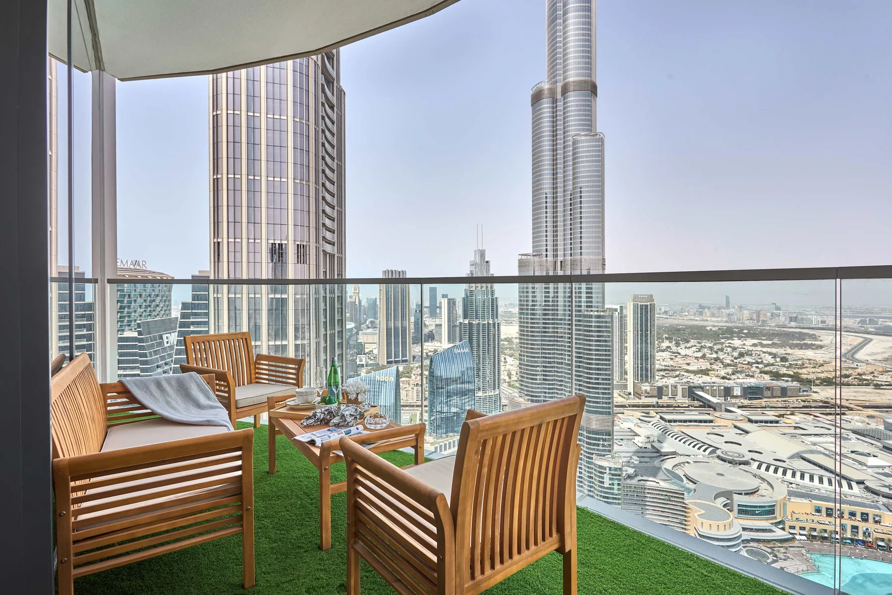 Una zona de estar en un balcón con muebles de madera y el horizonte urbano tras la barandilla de vidrio.