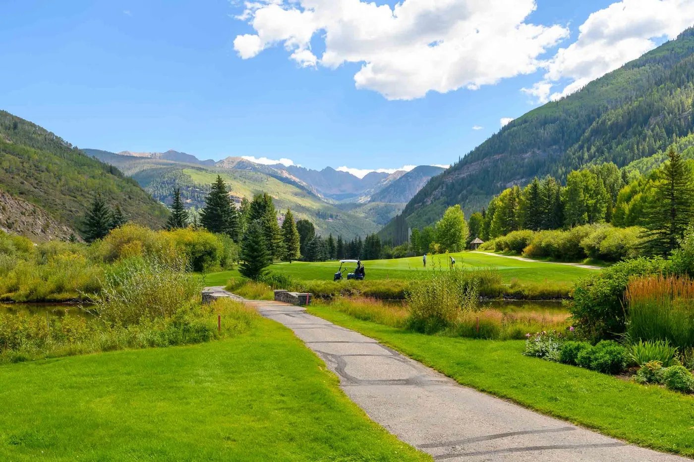 A paved path winds through a green valley with forested mountains beyond.