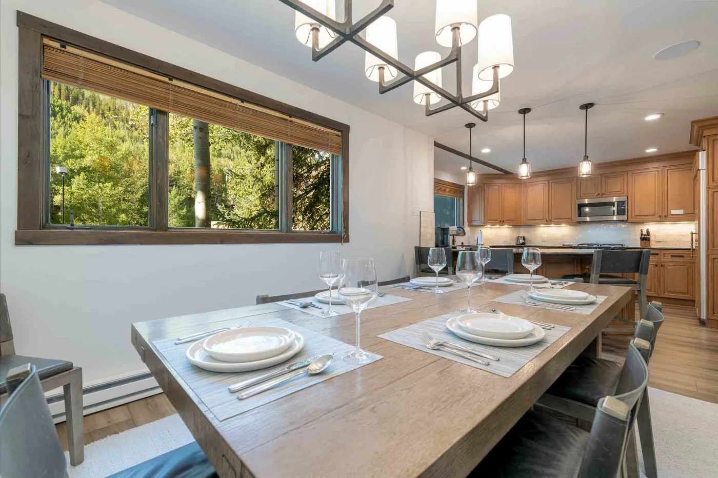A dining area with a long table set beside a large window, with a kitchen beyond.