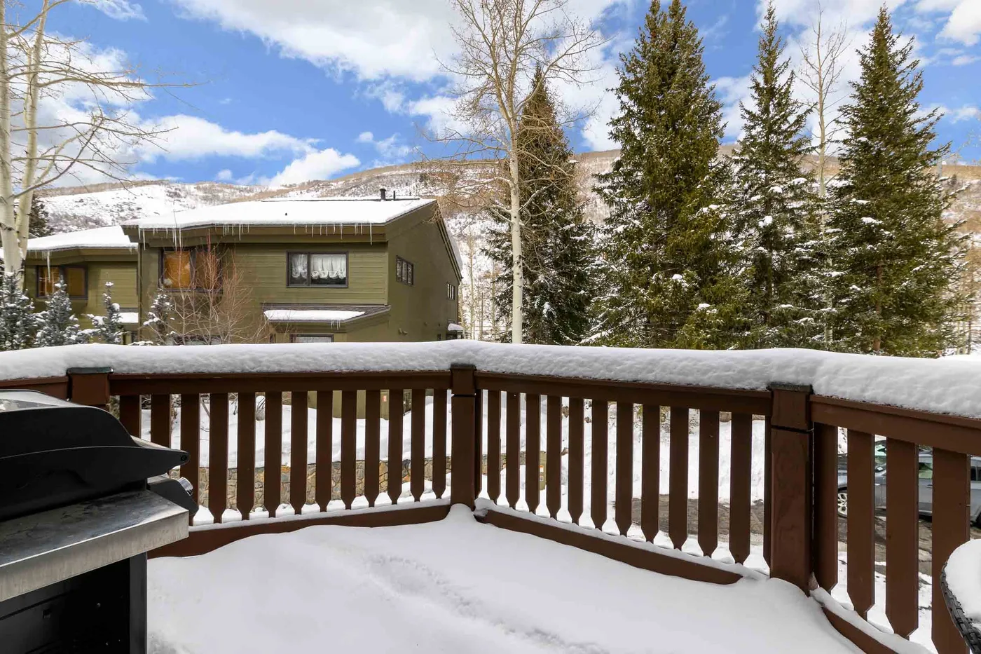 A snow-covered deck overlooks evergreen trees and a nearby building.