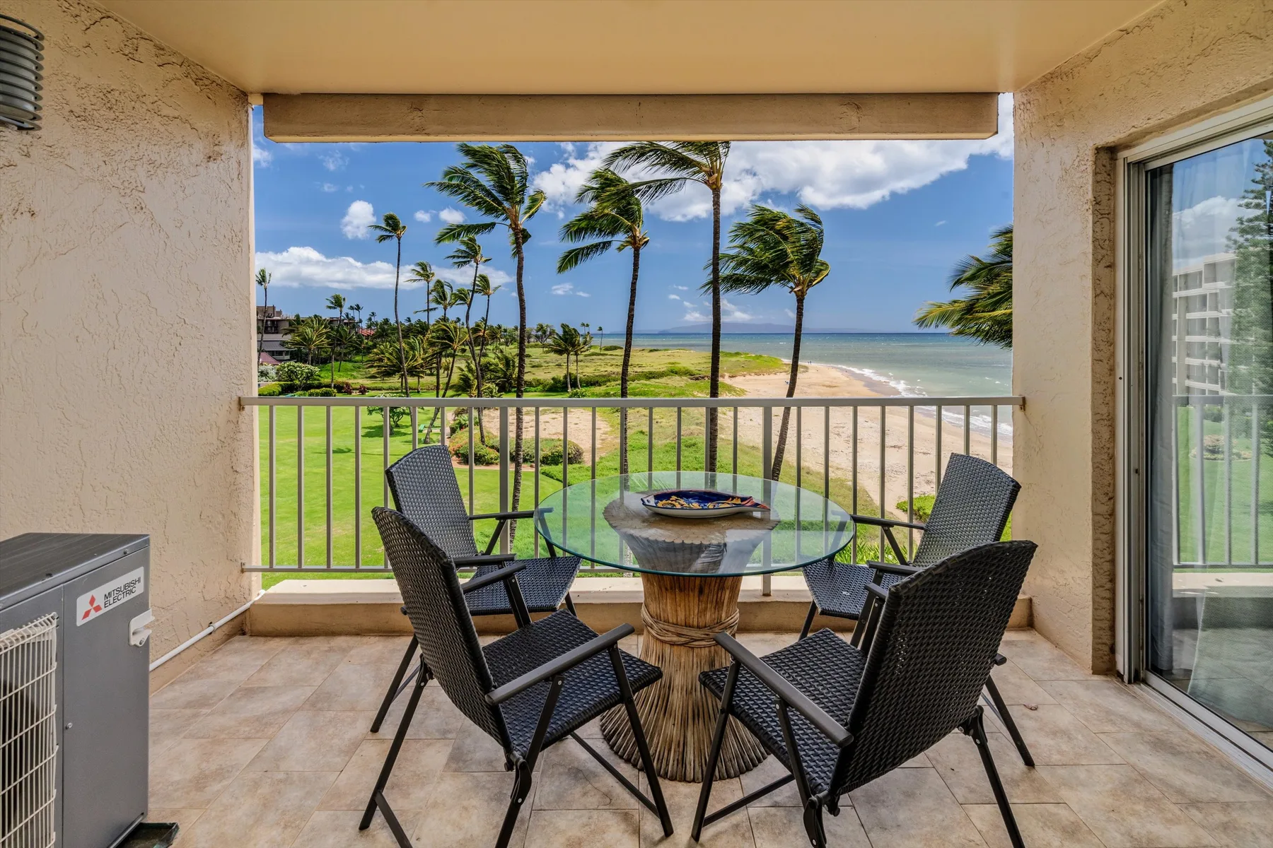 Un patio cubierto tiene una mesa de vidrio y asientos con vista a las palmeras y la playa.