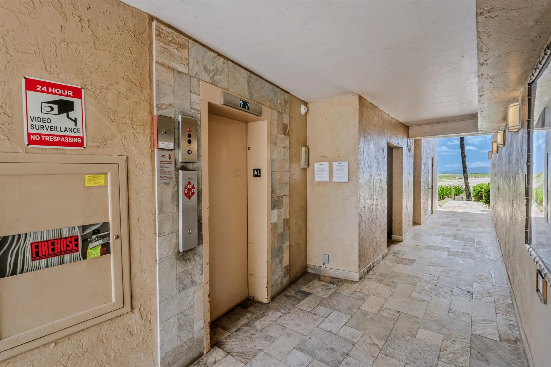 Un ascensor da a un pasillo con suelo de piedra y ventanas a un lado.