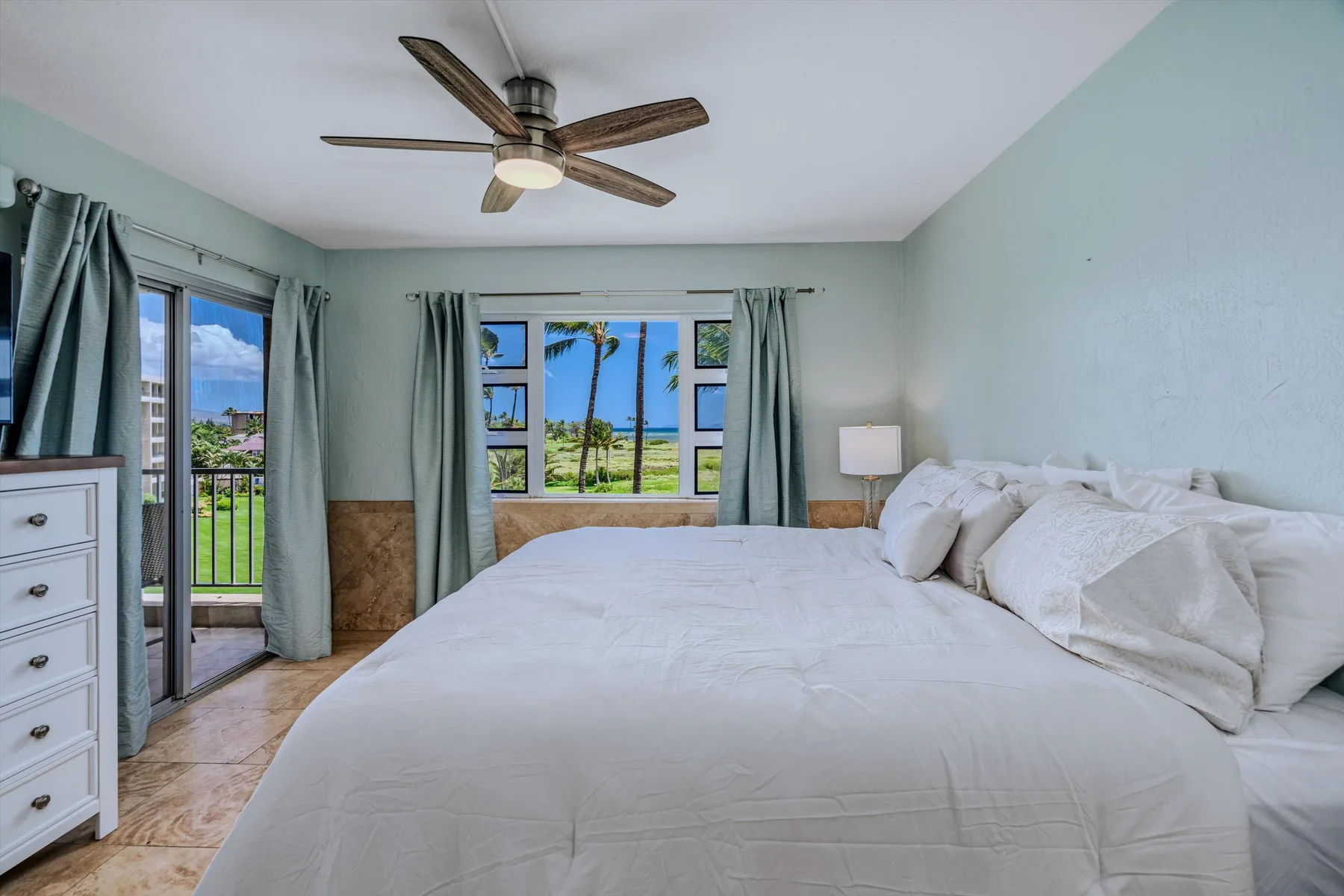 Un dormitorio luminoso con una cama grande y ventanas con vistas a palmeras.