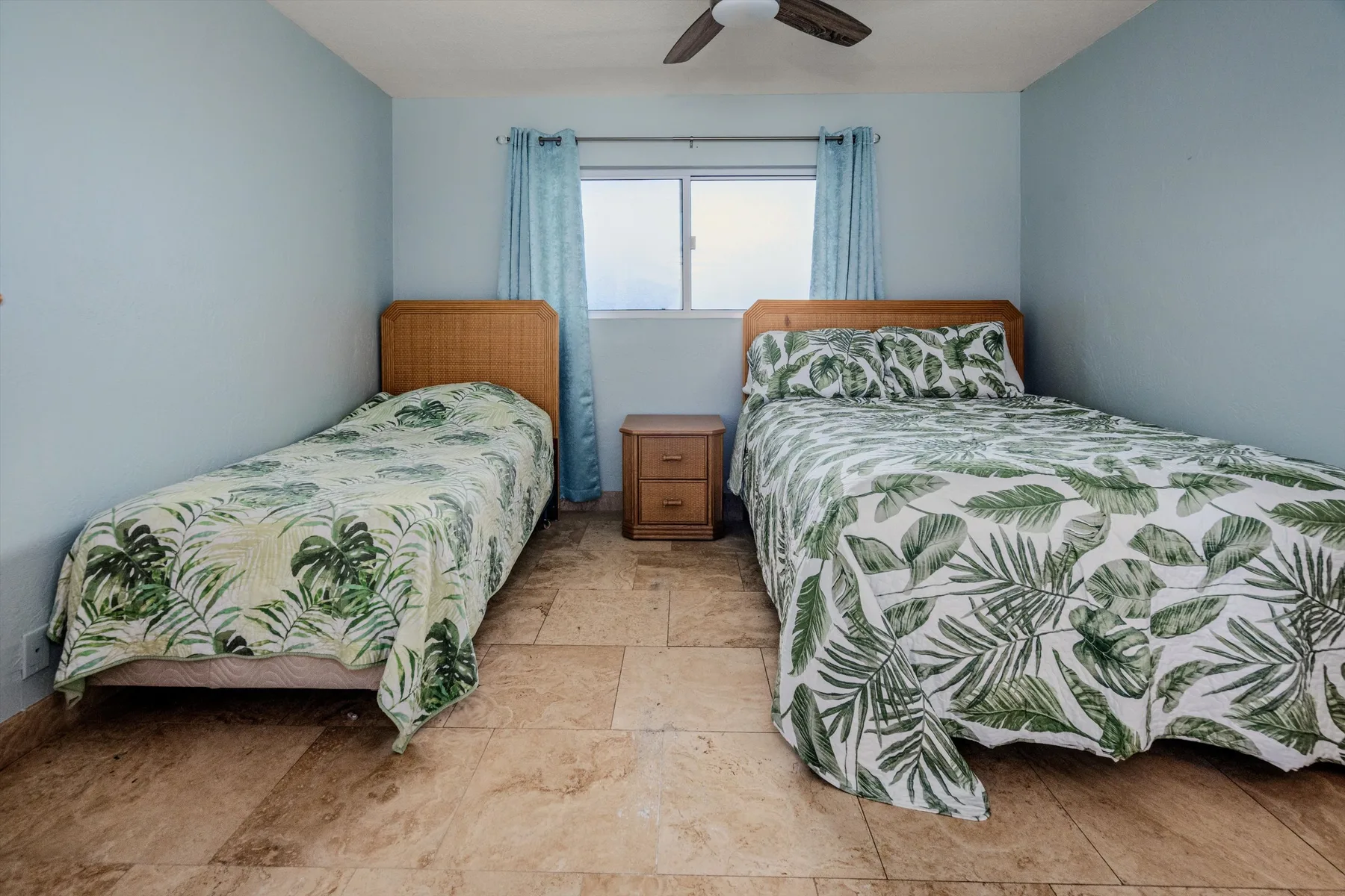 Un dormitorio con dos camas con ropa de cama estampada con hojas y una ventana entre ellas.