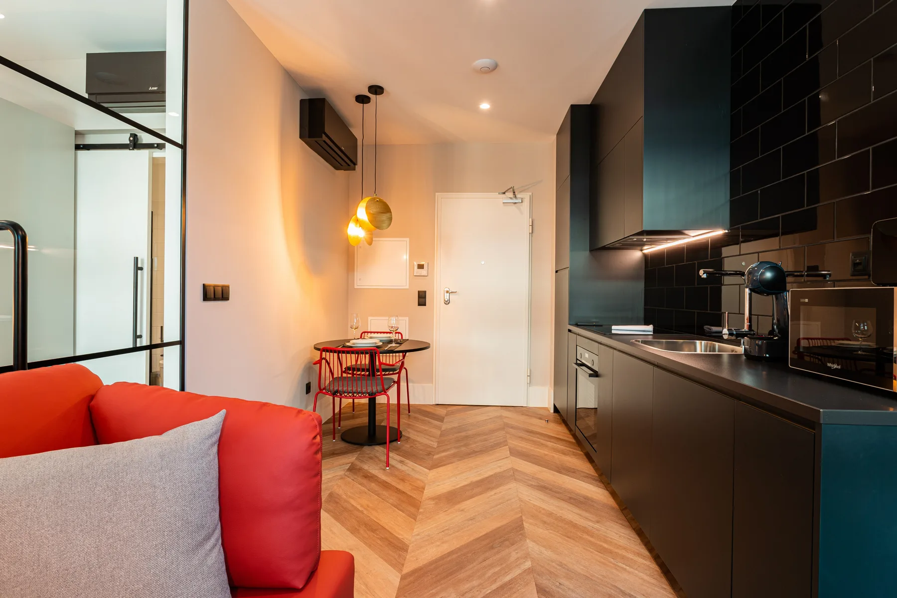 Un interior de apartamento combina un sofá rojo, una mesa de comedor y una cocina oscura.