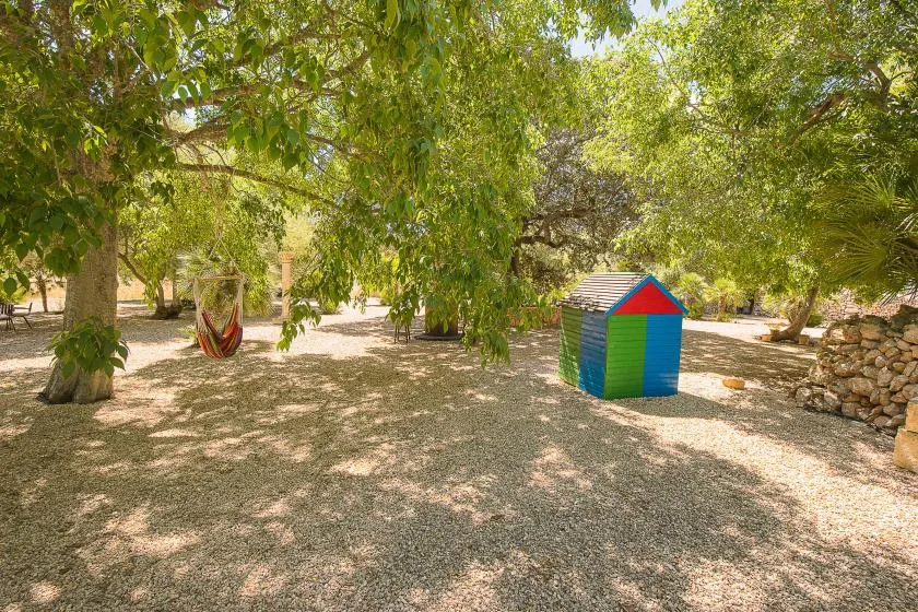 Un jardín sombreado con árboles frondosos, una hamaca y una pequeña casita de juegos colorida.
