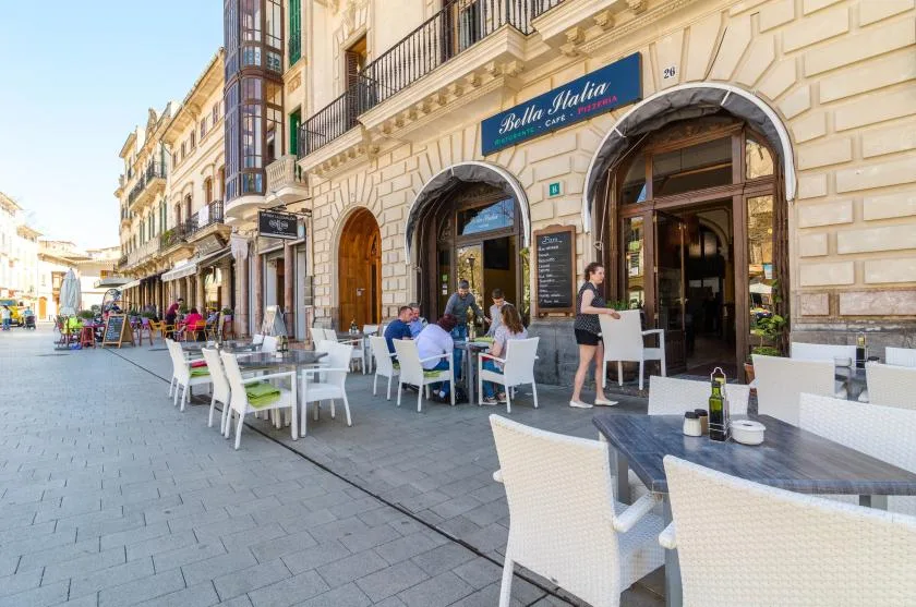 Mesas al aire libre de un café junto a la entrada arqueada de un edificio peatonal.