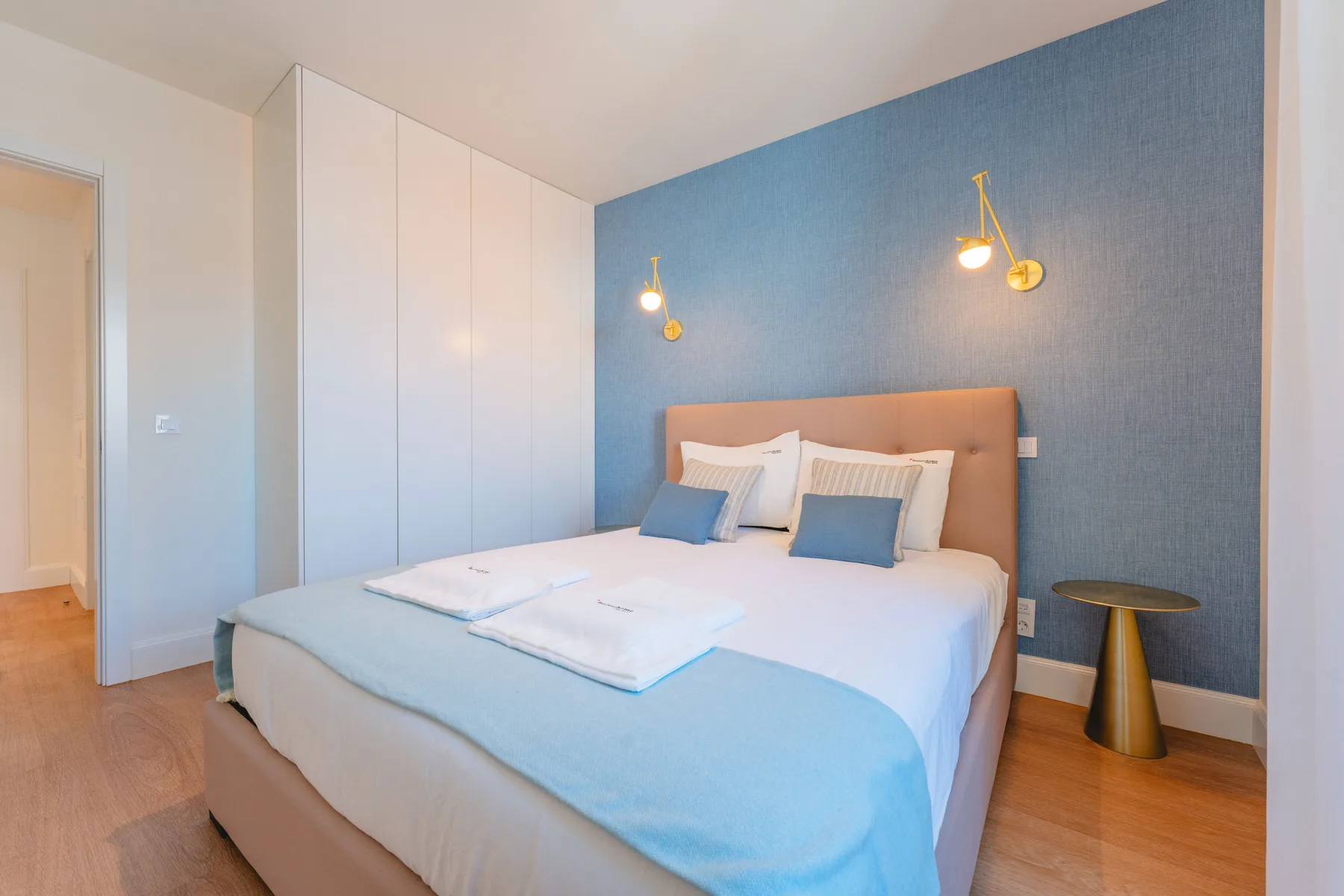 Un dormitorio con una cama vestida con ropa de cama blanca y azul frente a una pared azul.