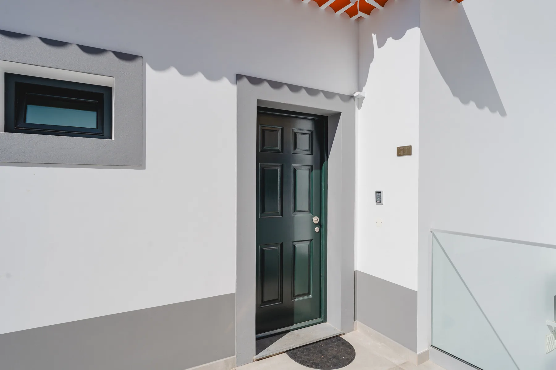Una puerta de entrada oscura está en una pared exterior blanca junto a una ventana pequeña.