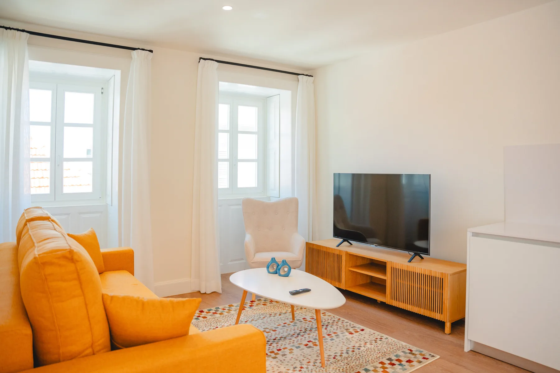 Un salón luminoso con asientos, un televisor y dos ventanas altas.