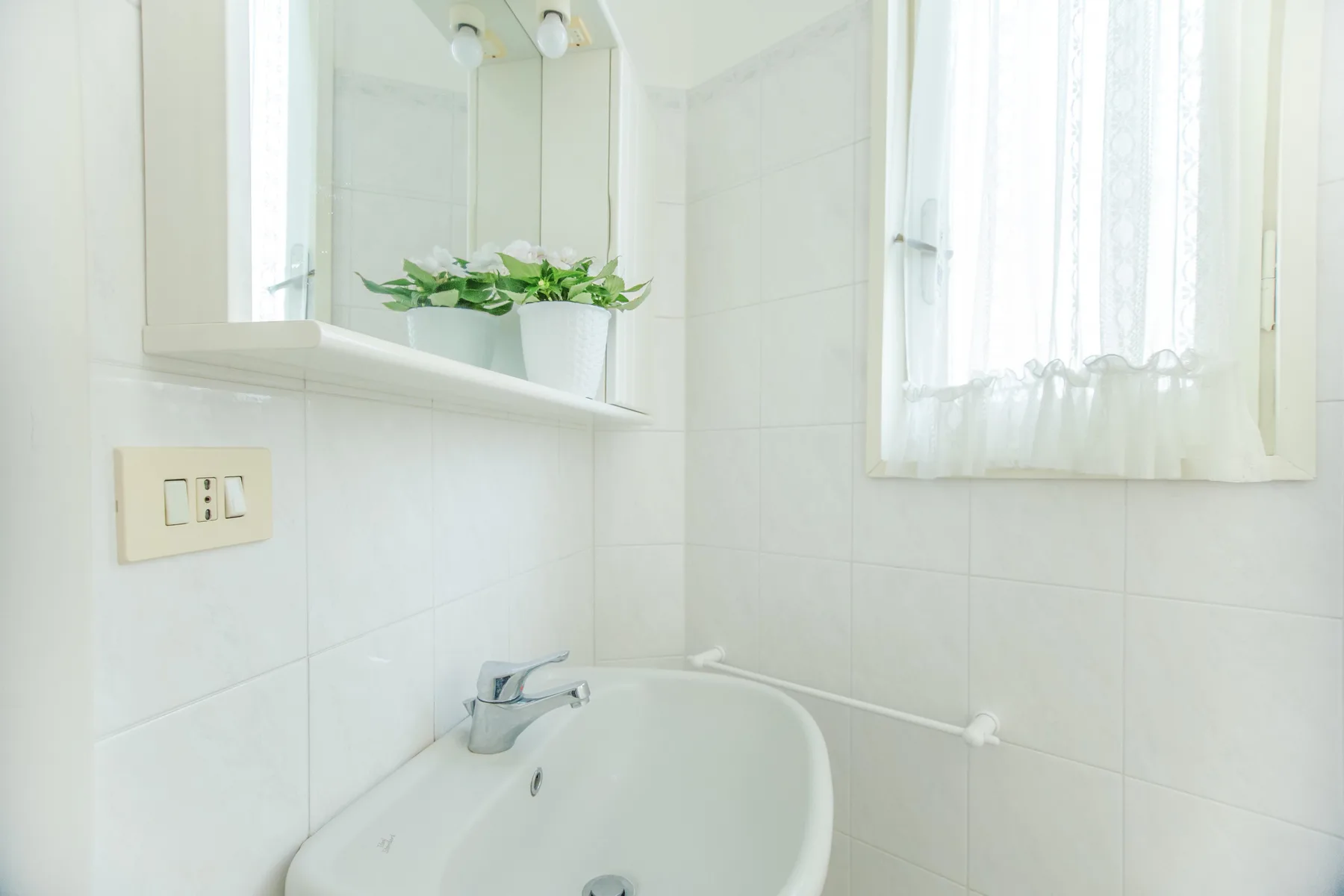 A bright tiled bathroom with a bathtub, sink, and curtained window.