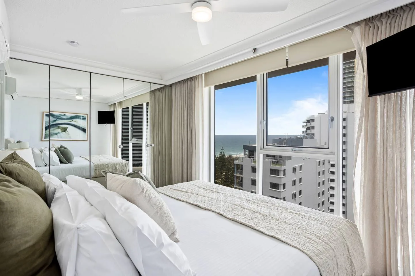 A bright bedroom with a bed beside large windows overlooking the ocean and nearby buildings.