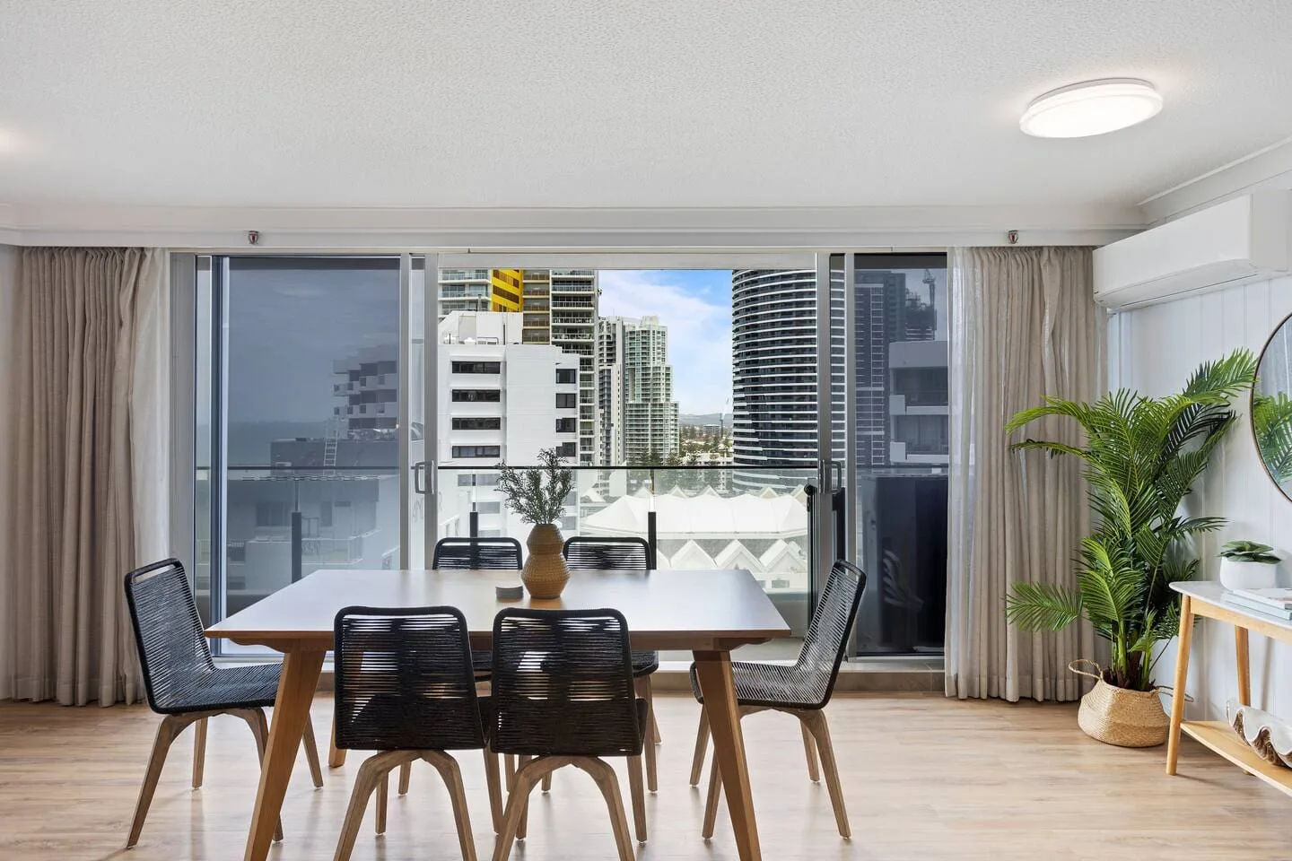 Un comedor tiene una mesa y sillas junto a grandes ventanales con vistas a edificios de la ciudad.