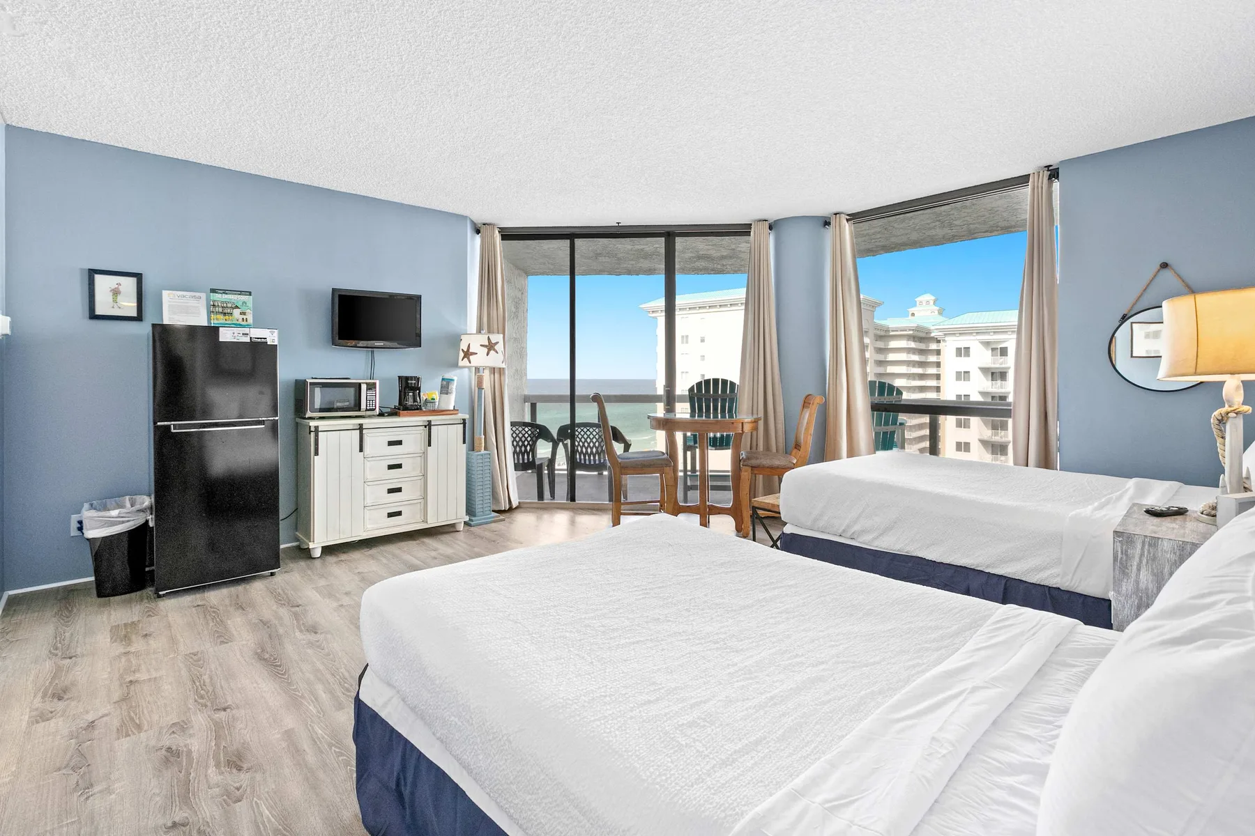 A bright bedroom with two beds, a small dining area, and large windows.