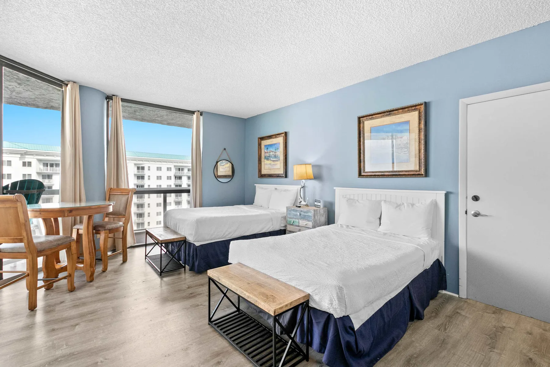 A bedroom with two beds, a small table, and large windows.