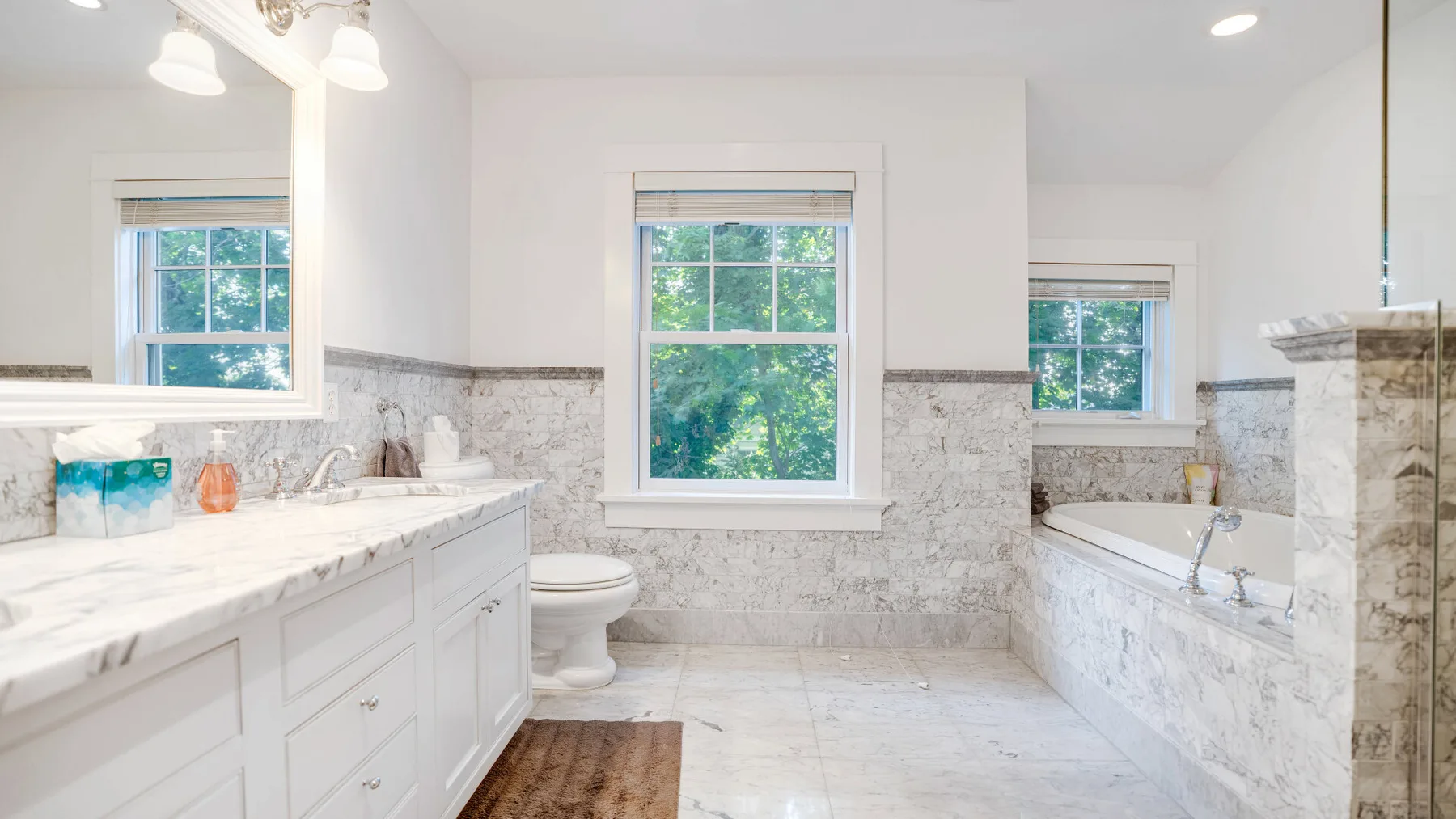 A bright bathroom with marble surfaces, a vanity, a bathtub, and a window.