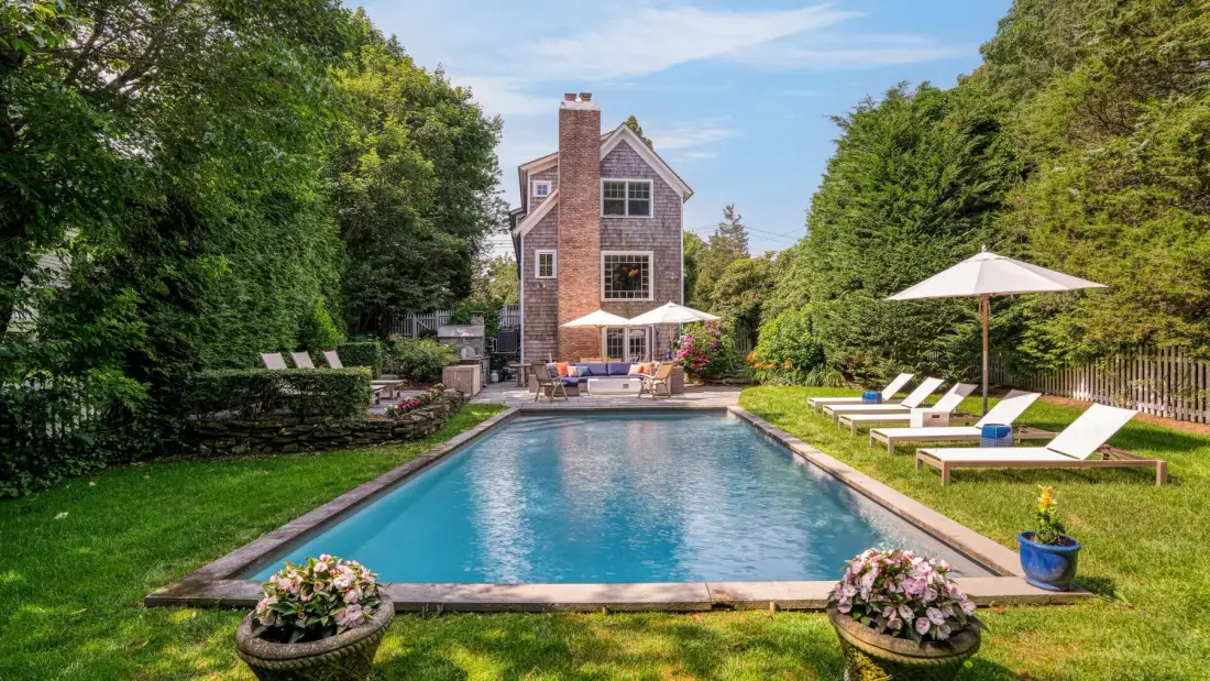 An outdoor swimming pool sits in front of a house, with lounge chairs on the lawn.
