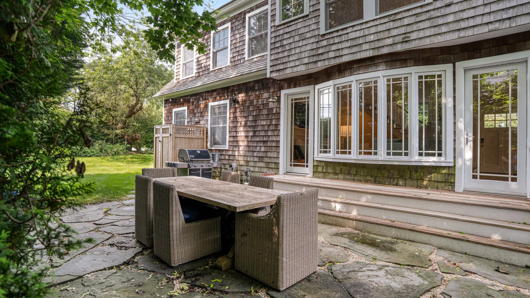 An outdoor dining area sits beside a shingled house and large windows.